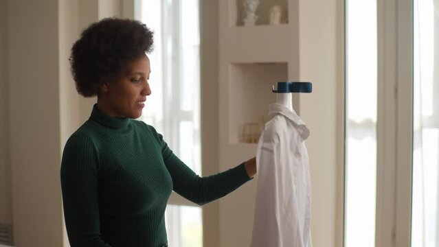 Tracking shot of cheerful African-American female ironing white shirt with manual garment steamer with positive expression. Black housewife using modern device standing at home indoors, slow motion.