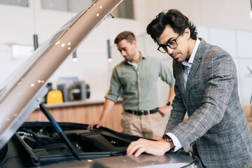 Fototapeta na wymiar Serious male client visiting dealership office and choosing new car, examine transport engine. Professional salesman describes customer about quality of vehicle in showroom. Concept of buying new auto