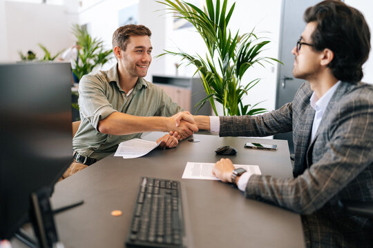 Side view of male real estate broker wearing business suit present and advise young man client on decision to sign insurance contract. Cheerful customer signing purchase agreement and shaking hands.