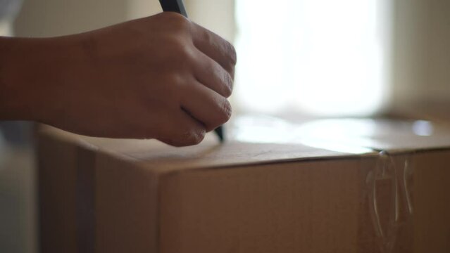 Close-up hands of unrecognizable African-American young woman writing label on cardboard box for moving out to new house sitting. Concept of new life in new home relocation. Shooting in slow motion.