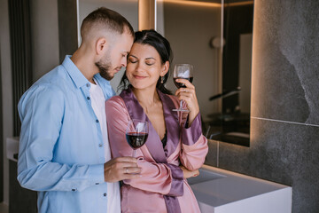 Young adult caucasian couple at home embracing holding glasses of wine, anniversary. Bearded man hugs wife at home, happy family. Happy woman in night gown eyes closed clings to husband.