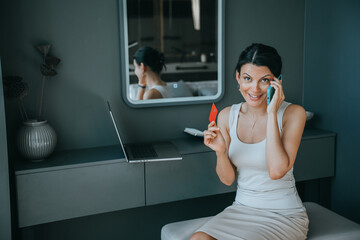 Fototapeta na wymiar Cheerful caucasian businesswoman in casual sitting on couch talks by phone looks at camera shows credit card. Smiley housewife making online purchase, talks with bank employee.