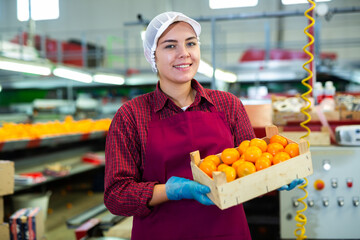 Smiling young female worker in fruit sorting and packing warehouse holding small wooden box of sorted ripe mandarins © JackF