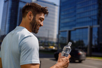 Smiling sport man holding water bottle after running in the city skyscrapers background