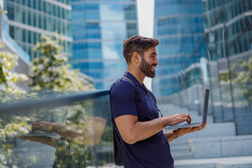 Back view of smiling male entrepreneur working on laptop standing on skyscrapers background 