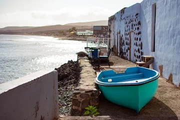 Papier Peint photo Lavable les îles Canaries Small green and blue fishing boat, on the ground next to a white house next to the shore of the beach at sunset.