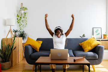 Happy african student woman sitting on sofa with laptop and celebrating success after reading unexpected message on e-mail