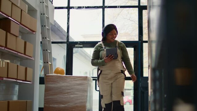 Relaxed african american warehouse picker listening music while checking labels on cardboard boxes. Smiling worker wearing heaphones and bopping head while walking through storage area aisles