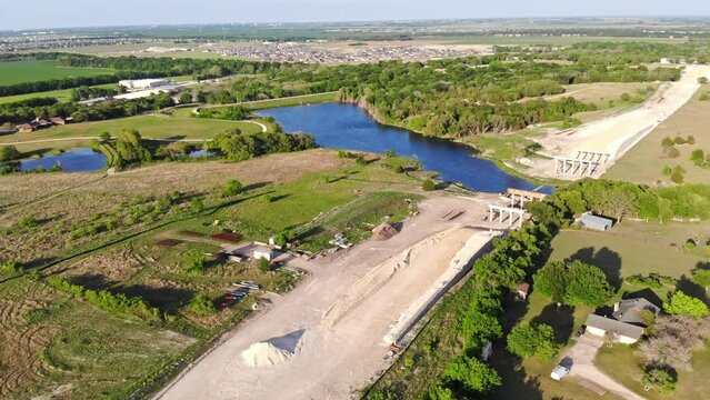 Flying down towards construction of a major road project which requires a bridge to cross a water reservoir. Homes with acreage on either side of the new road and a large sub-division in the distance