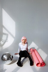 A young woman with pink hair sits elegantly in a minimalist studio, wearing a white blouse, black...