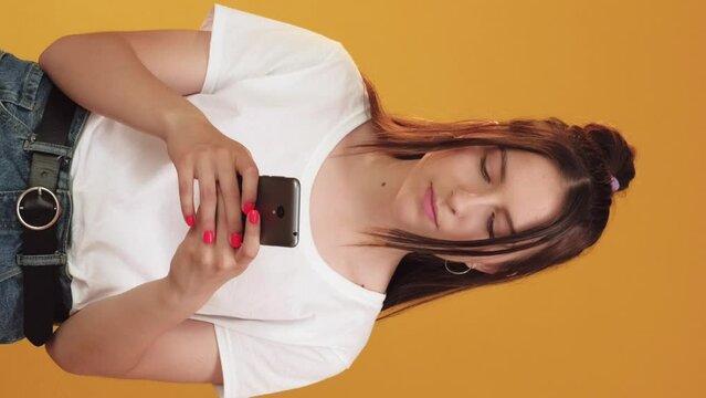 Vertical video. Mobile chatting. Text message. Pleased smiling woman typing in modern smartphone social media application isolated on orange background.