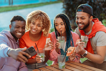 Group of smiling stylish African American  friends taking selfie, eating pizza, drinking lemonade sitting together in modern cafe  - obrazy, fototapety, plakaty