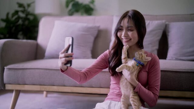 Happy Asian Woman taking selfie photo with smartphone camera of herself and her cat on couch at home.