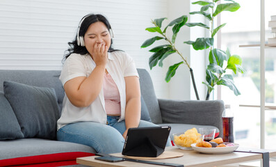 Asian young overweight oversized fat chubby plump unhealthy female teenager in casual outfit sitting drinking cola soft drink eating junk fastfood chips donuts when watching tablet in living room - obrazy, fototapety, plakaty