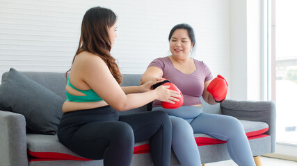 Obraz premium Two Asian young overweight oversized fat chubby plump female sportswomen in casual sportswear sitting on cozy sofa wearing boxing gloves ready for exercising training workout in living room home gym