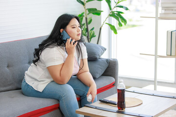 Asian young chubby fat unhealthy oversized overweight lazy female teenager in casual outfit sitting on cozy sofa on call via smartphone holding reading loss weight pill label in living room at home