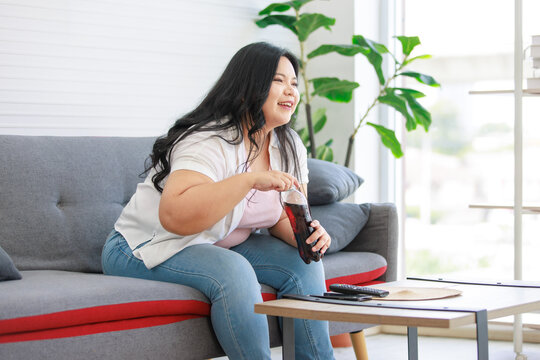 Asian young chubby fat unhealthy oversized lazy female teenager in casual outfit sitting on cozy sofa drinking cola soft drink from plastic bottle watching television show in living room at home
