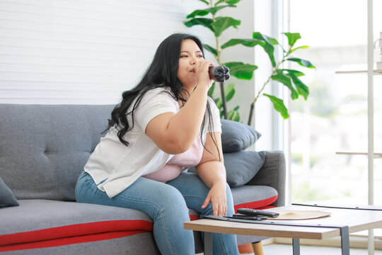 Asian young chubby fat unhealthy oversized lazy female teenager in casual outfit sitting on cozy sofa drinking cola soft drink from plastic bottle watching television show in living room at home