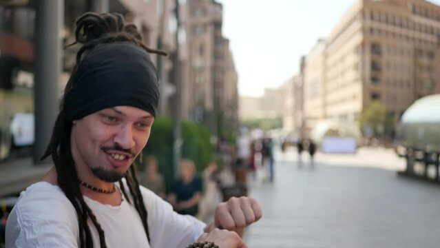 Portrait of a dancing man with a bun of long dreadlocks and a beard smiles happily and makes rhythmic movements outdoors in the city among passing people. Interesting stylish man