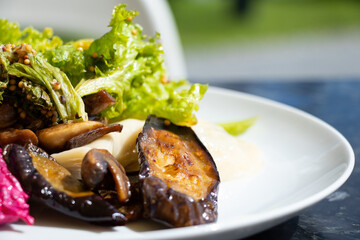 Cabbage leaves and mushrooms and a slice of fried eggplant on a white plate in the sun on the table, cooked vegetables for lunch