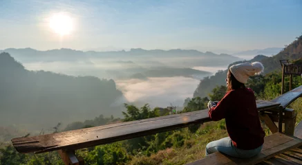 Schilderijen op glas Asian female tourist is sitting and enjoying the morning sunrise and mist on the mountain. © pakorn