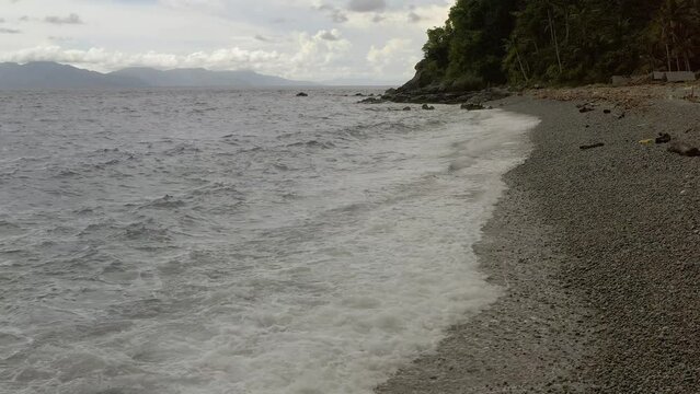 Coastal Dolly Shot Along the Pebbled Beach with Ocean Waves with Scenic Mountainous Background in Looc Bay, Philippines.