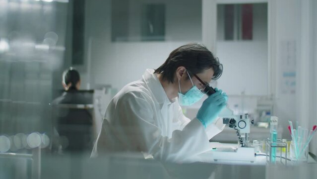 Side view of Asian scientist wearing protective mask and gloves sitting at workplace, using microscope and writing down results of research while working in laboratory