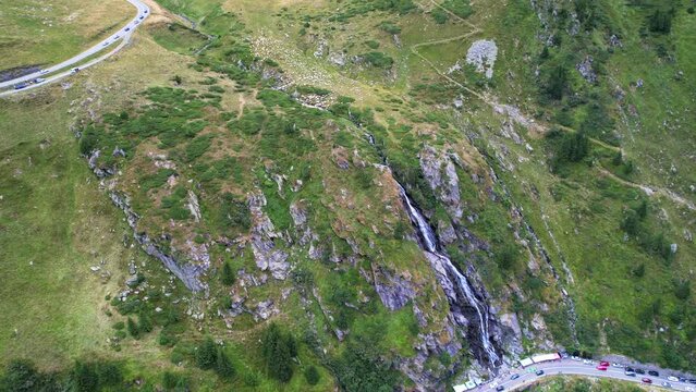 Cascading waterfall cuts rocky slope at pull off on Transfagarasan Serpentine Road, aerial panoramic pedestal