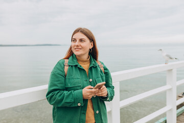 Beautiful young smiling woman with backpack using smartphone during walk at the beach. Hipster girl with backpack hold on smart phone gadget outdoors