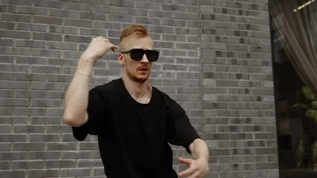 A dancer in black glasses dances electro against the background of a gray brick wall.