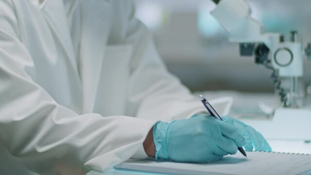 Tilt down shot of Asian scientist in lab coat and gloves writing down notes and taking test tube while working at desk in laboratory