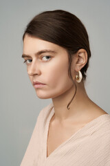 Close-up shot of a young European woman without makeup wearing stud earrings decorated with pearl beads. A girl with gathered hair and pearl earrings is on a gray background. Front view.