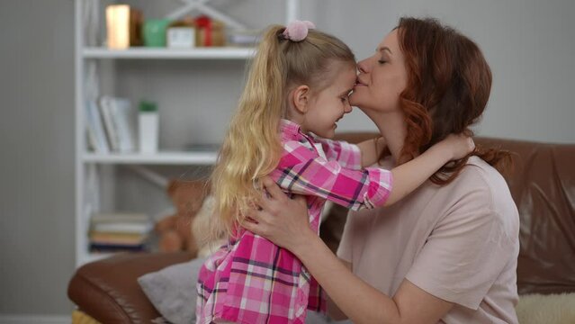 Side view loving mother rubbing noses with daughter kissing kid forehead hugging child. Happy smiling confident Caucasian woman and girl embracing at home indoors