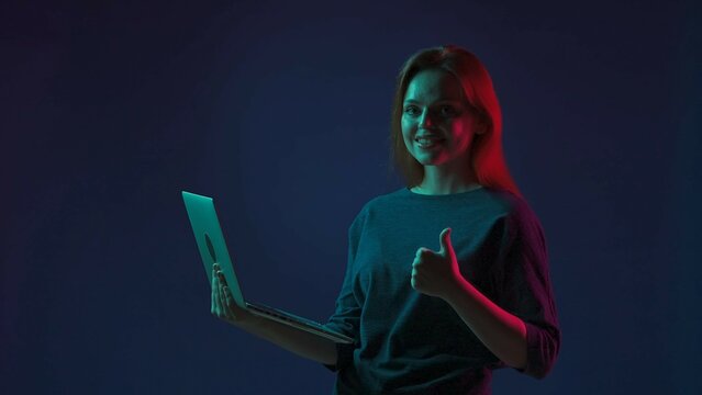 Woman showing thumbs up while using laptop. Woman with laptop in hand in studio on blue background in pink and green neon light. Remote work, freelancer.