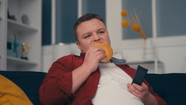 Lazy young man eating junk food and watching tv on couch, unhealthy lifestyle
