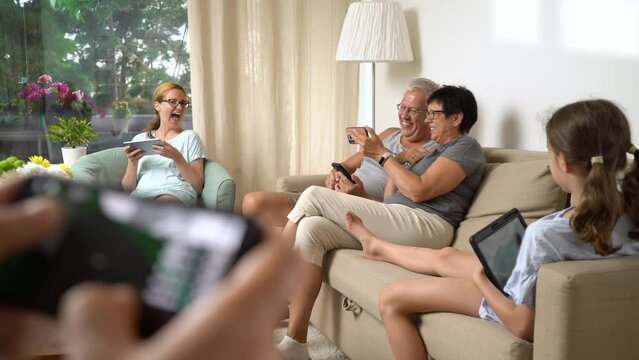 Big happy family is playing online mobile game together at home, people different ages having fun sitting on sofa. Parents, grandparents and little girl relaxing in living room with mobile devices