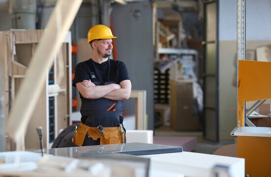 Portrait of young attractive man in work clothes and yellow helmet crossed arms smiling at shop for manufacturing furniture and details home interior