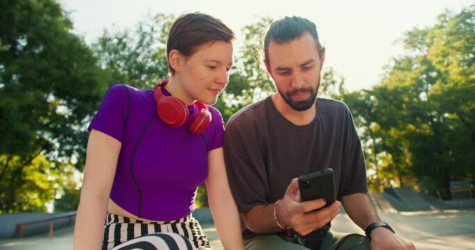 A brunette guy in a gray t-shirt shows a girl with a short haircut in a purple top and red headphones something on his black phone in a skatepark in summer