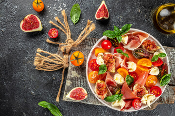 Homemade salad with mozzarella, figs, prosciutto, fresh basil leaves and tomato on a dark...