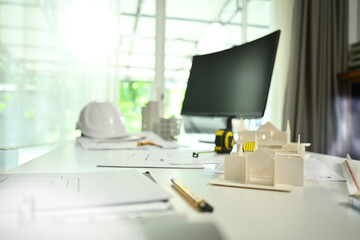 Architect desk with house model, blueprints, safety helmet and computer monitor on white table