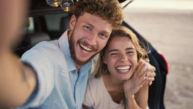 POV: A bearded guy with curly hair in a blue shirt is smiling at the camera with his blonde girlfriend while waving takes a selfie in the trunk of a black car, decorated with light bulbs