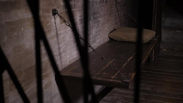 An old prison cell with a wooden bed and a pillow, a metal grille in the foreground. A dark old prison cell with a brick wall.