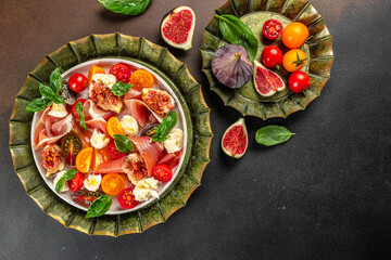 delicious figs and prosciutto salad, mozzarella, fresh basil leaves and tomato on a dark...