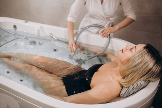 Pleases spa centre employee putting a flexible pipe into water in a bath with a client for hydromassage