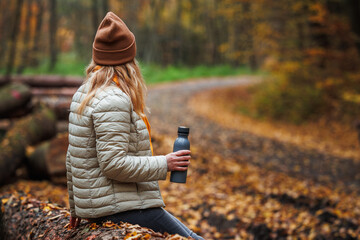 Woman sitting and resting on log in autumn woodland. Hiker holding thermos flask with hot drink....