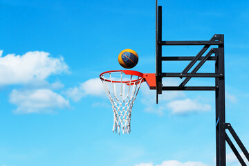 Basketball basket and ball against a background of blue sky and clouds. Sports equipment in an open area. Copy space