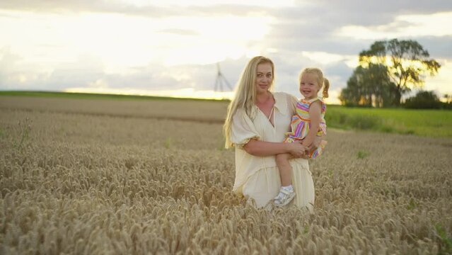A woman with a little girl in her arms stands looking at the camera and smiling. Beautiful background with wheat field and sky at sunset. High quality 4k footage