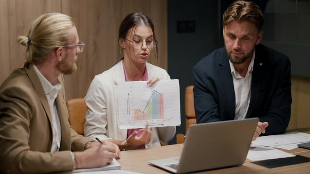 A brunette girl in a white suit in a pink shirt and round glasses shows a graph on a sheet of paper to her interlocutor on a video conference in the company of two guys in business suits in a modern