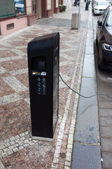 An electric charging station in use on a cobbled street. A black charging station with the cable connected to a parked black car.