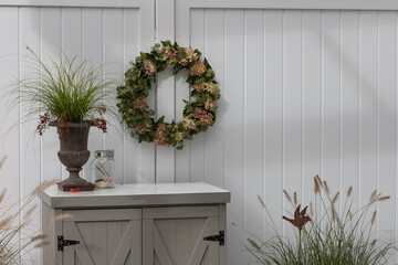 A little cabinet on a terrace in front of a white divider with decorations like a flower wreath, an...